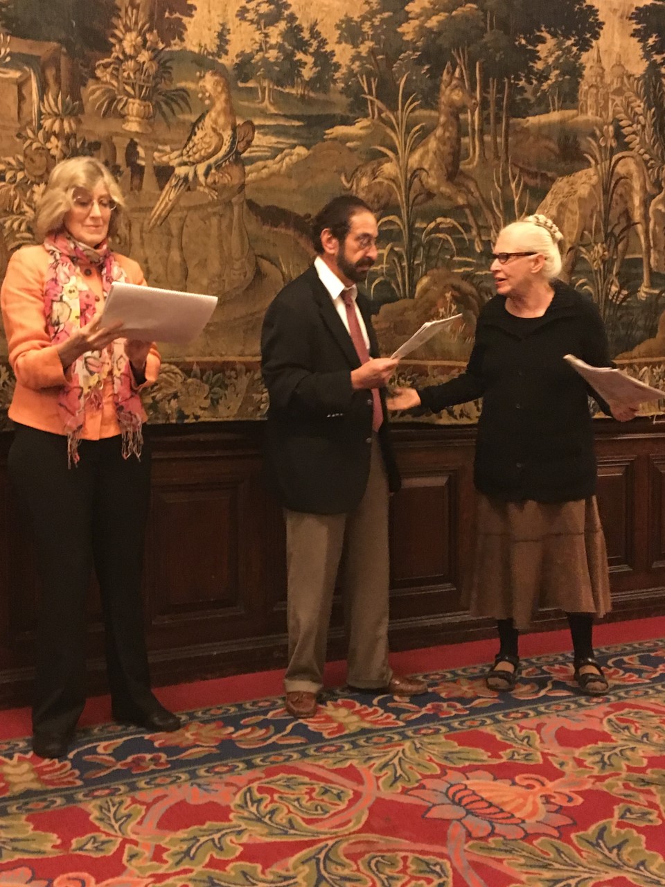 Three people stand in an ornatly-decorated room in formal clothes reading from scripts dramatically.